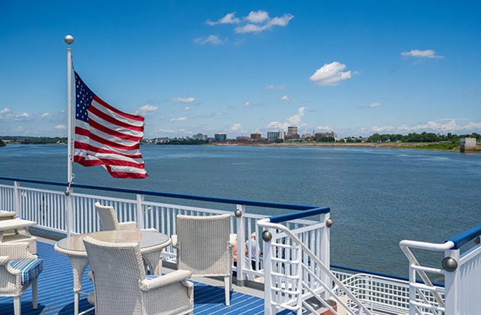 On the deck of an American Riverboat cruising on the Ohio River, there is a round, white wicker table with a glass top and white wicker chairs surrounding it. To the left, there is a large American flag on a flag pole blowing in the wind with shoreline in the distance.