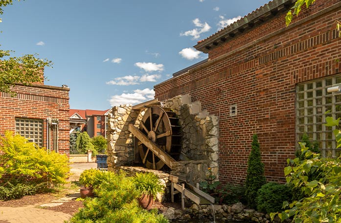 A rustic, old wooden water wheel is set in the corner of two stone walls with rugged edges against a brick building near the Mississippi River in La Crosse, Wisconsin. There are several bushes, shrubs, and potted plants, and a separate corner of a brick building to the left.