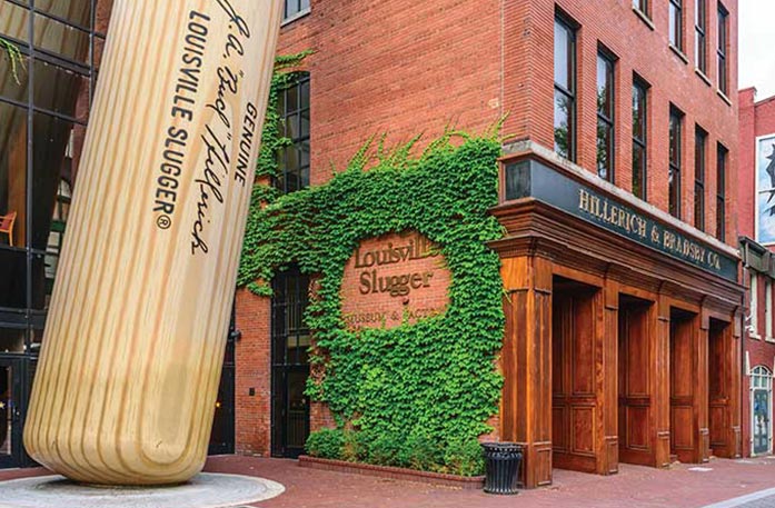 In front of the Louisville Slugger Museum and Factory, a giant wooden Louisville Slugger baseball bat leans against a red brick building, Hillerich & Bradsby Co., with "Louisville Slugger" in gold writing on the left side of the building surrounded by bright green vines.