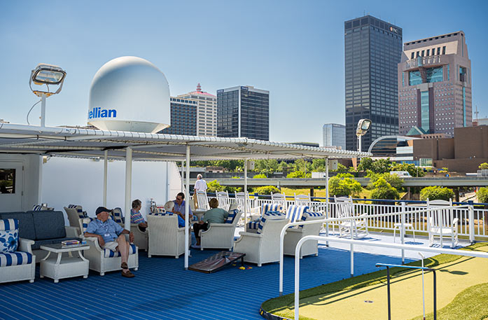 A few cruise guests sit in white patio chairs with blue and white striped pillows on the American Riverboat Sun Deck under a sun shade that has tow large spotlights on each end. On the right of the deck, there is a putting green hole, and a corn hole board to the left of it. In the background, there are several skyscrapers.