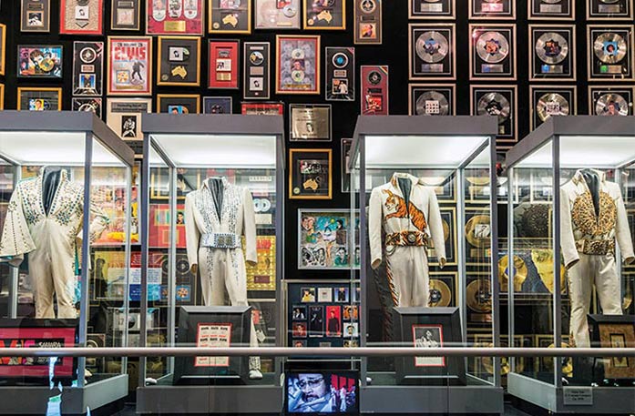 An Elvis Presley exhibit at Graceland in Memphis, with several of his iconic white outfits displayed in glass cases with a wall filled with his records and posters.