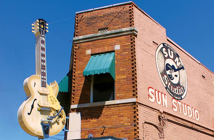 Sun Studio, a brick building with a green awning over the window with a large mural of a record with a guitar in the middle and the words "Sun Studio" in white on and underneath it. There is a very large light tan colored guitar sculpture to the left of the building.