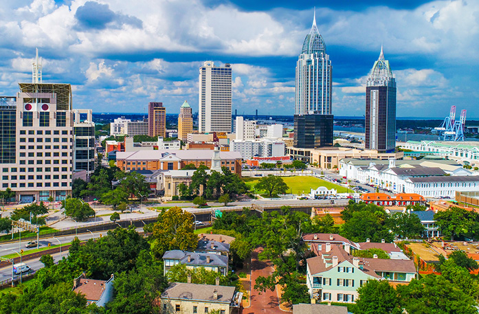 An aerial view of the Mobile cityscape, where there are several buildings and skyscrapers under a cloudy sky, including RSA Battle House Tower, a large skyscraper with a rounded top and sharp spire, and Mobile Government Plaza, a high-rise building with large solar panels on the roof. There is a highway in the center that divides the two sides of the city.