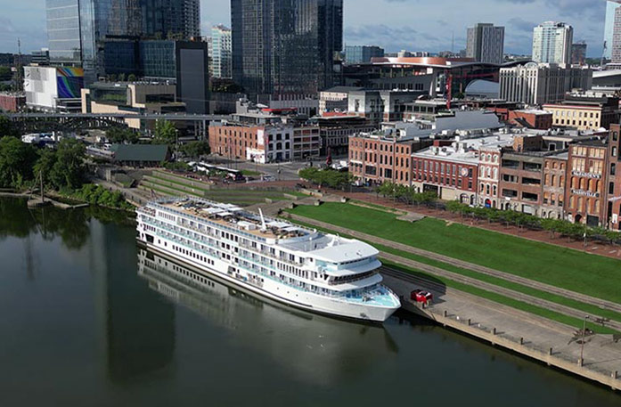 The American Riverboat American Serenade docked on the Cumberland River with an aerial view of Nashville's city landscape, including brick buildings and tall skyscrapers.