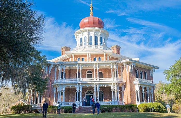 Longwood, a large, octagonal brick mansion in Natchez with a red byzantine onion-shaped dome with ornate white porches, surrounded by trees, under a blue sky. People are walking in the front yard. 