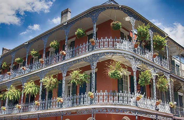 An ornate red-brick building with two-story wrought-iron balconies adorned with hanging plants under a blue sky in the historic French Quarter in New Orleans. 