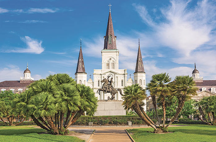 The St. Louis Cathedral with three spires stands in Jackson Square in New Orleans, while a statue of a rider on a horse is centrally placed in a manicured park with trees and bright greenery.