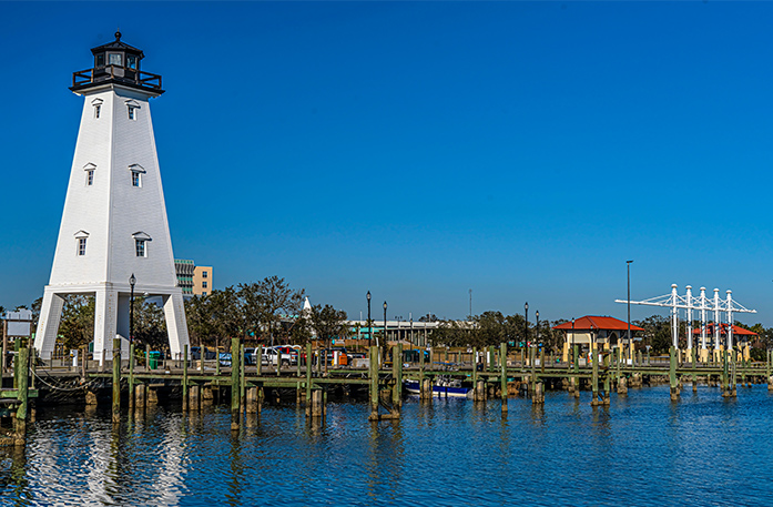 A wide view of Ocean Springs, with Ship Island Lighthouse, a tall white lighthouse with an open bottom, on the left side of the dock on the water. 