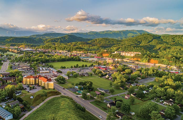 An aerial view of buildings, open green flatland, and the Great Smoky Mountains in the distance in Pigeon Forge. The buildings have a bright orange glow from the sunrise that is out of view.