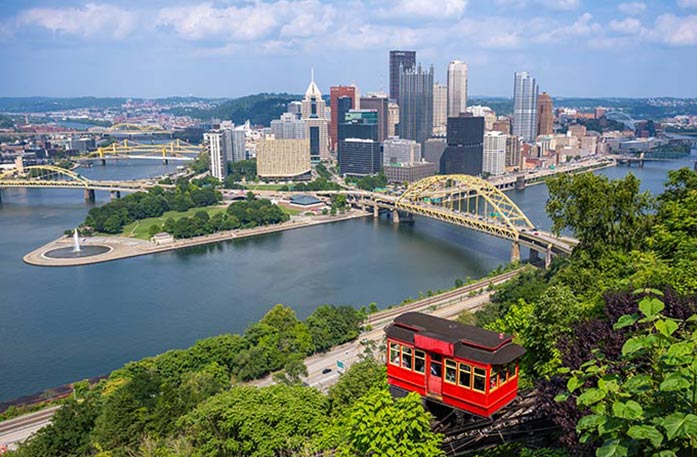 A red cable car descends a forest hill overlooking the Ohio River and a Pittsburgh cityscape featuring a brideg with a yellow truss in the middle and  several skyscrapers under a partly cloudy sky.