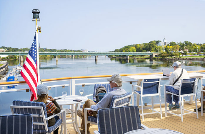 A couple sits at a table beside an American flag on an American Cruise Lines Riverboat's sun deck looking out at the Mississippi River, while a man sits to the right of them typing on his computer.