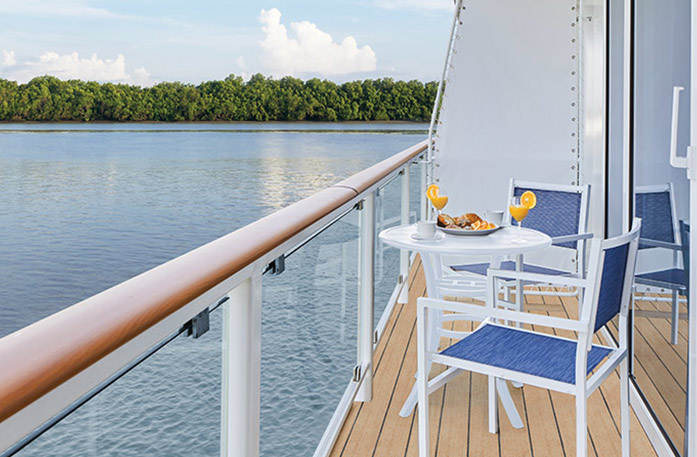 On an American Riverboat private balcony, two blue and white chairs surround a round table with a plate of food and two yellow drinks with large lemons on the rim on the balcony of one of the ships staterooms.