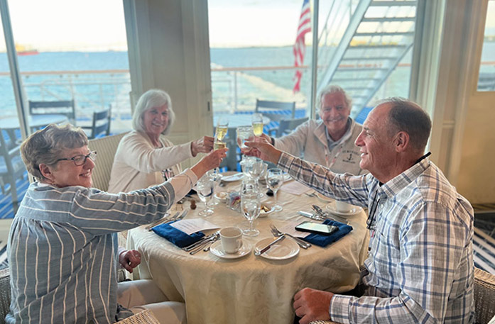 Two couples sit together at a round dinner table on an American Riverboat having a toast with their champagne glasses before a meal served by American Cruise Lines.