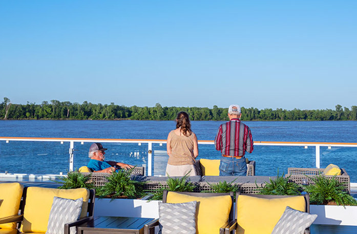 On the American Riverboat Sun Deck, a couple is standing against the glass railing gazing at the Mississippi River, the trees lining the horizon line, and the clear blue sky. There are four patio chairs with yellow and grey pillows in the foreground, and in between those and the couple, there is a man sitting on a chair facing the right.