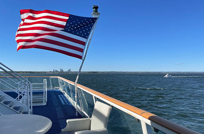 On the deck of the American Heritage paddlewheeler, there is a large American flag, with it's pole on the glass railing, blowing in the wind with the views of the Mississippi River. The floor of the deck is dark blue, a few stairs can be seen to the left, and there is an empty tan colored chair and table in the foreground.