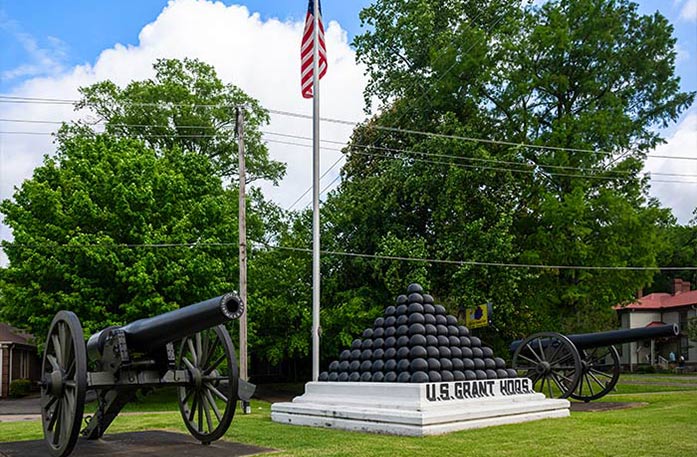 A memorial structure with a pyramid of black cannonballs on top sits between two black cannons at Shiloh National Military Park in Savannah, with an American flag on a flag pole in the background among full, green trees.