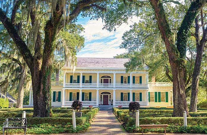 The historic St. Francisville landmark the Rosedown, a yellow house with bright green shutters and two-story white pillars with a brick pathway surrounded by perfectly trimmed bushes and large trees leading to the red door. 