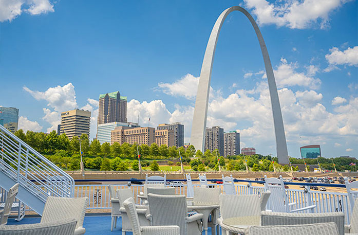 Multiple tables and chairs line the blue deck floor of an American Cruise Lines American Riverboat with the view of the St. Louis Gateway Arch and several skyscrapers in the background.