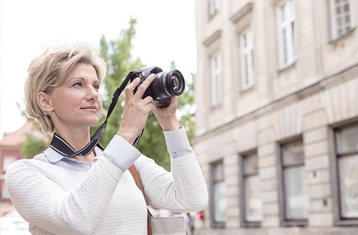 A woman with short blonde hair wearing a white sweater holds a black camera, taking a photograph of a sight out of view.