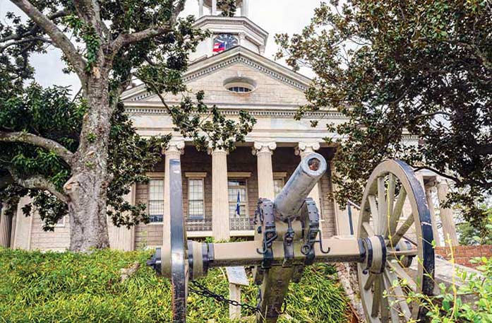 A cannon with two large wheels is pictured in front of the historic Old Warren County Courthouse, a light brick building with pillars out front in Vicksburg.