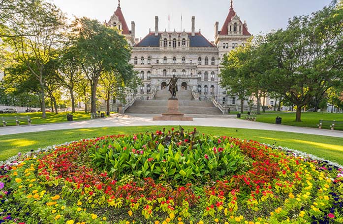 The New York State Capitol, a large building with two red spires on each side and a bronze statue in front of a wide staircase along a circular pathway. In the foreground, there is a circular garden with evenly placed yellow, red, pink, and green flowers and plants.