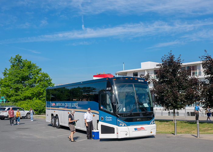 An American Cruise Lines coach bus, with the company name and American flag logo on the side, parked in front of a docked Coastal Cat in Albany. There are a few guests approaching the bus and the bus driver standing beside the open door.