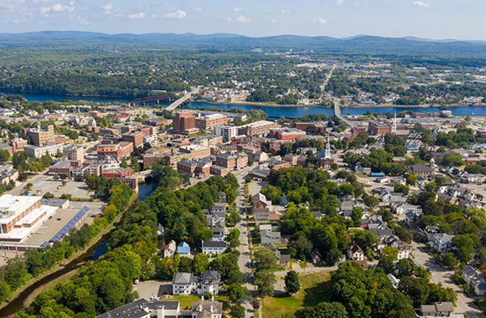 An aerial view of Bangor, with countless buildings and trees split between  the Penobscot River in the center. There are small mountains on the horizon line in the distance.