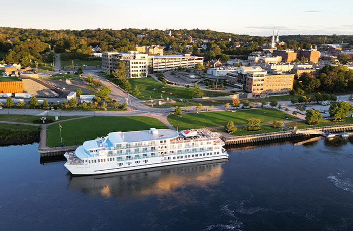 An aerial view of an American Cruise Lines small cruise ship docked in Bangor, with buildings and thick trees in the distance on shore.