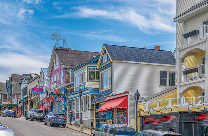 A roadside view of a line of buildings in Downtown Bar Harbor, with several cars parked alongside the sidewalk. One of the buildings is pink, one is yellow, and the others are a mixture of blue and tan.