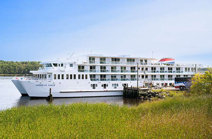 American Eagle Coastal Cat docked in Bath. There is tall grass in the foreground, and an American Cruise Lines tour bus beside the small cruise ship.