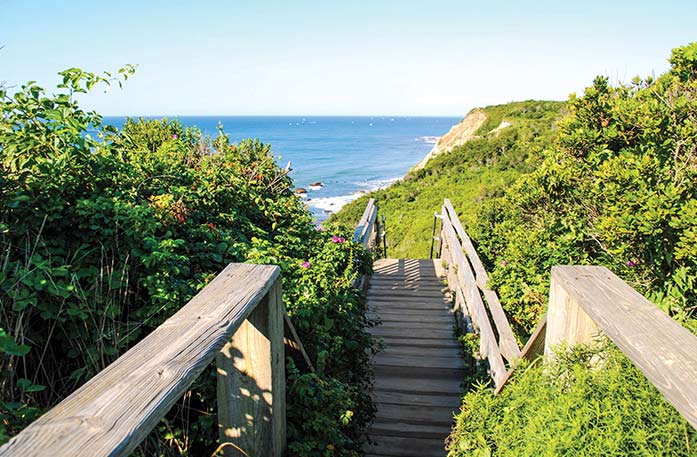 A wooden bridge in Block Island that allows people to walk down steps to the ocean. Each side of the bridge has thick wooden railings and is surrounded by greenery.