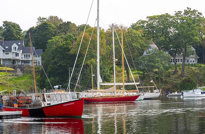 A red boat is docked in Boothbay Harbor, alongside several red and white sailboats beside a grassy hill that leads up to two large houses.