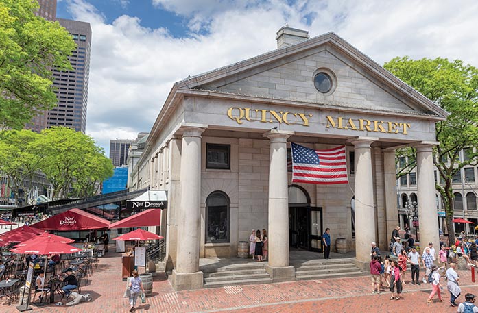 Quincy Market, a light brick building in Boston with four pillars in the front and an American flag between the two in the middle, and several chairs and tables with red umbrellas to the left of the building. A gold sign with "Quincy Market" is above the entryway, and there are several people walking outside the building. 