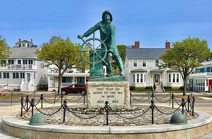 Gloucester Fisherman's Memorial, a large, green statue of a fisherman standing and holding onto the wheel of a ship in Gloucester, with a large stone stand below it with the phrase "They that go down to the sea in ships" and the years "1623" and "1923" on it. There is a circular platform made of stone with black railings surrounding the statue, and four houses behind it. 