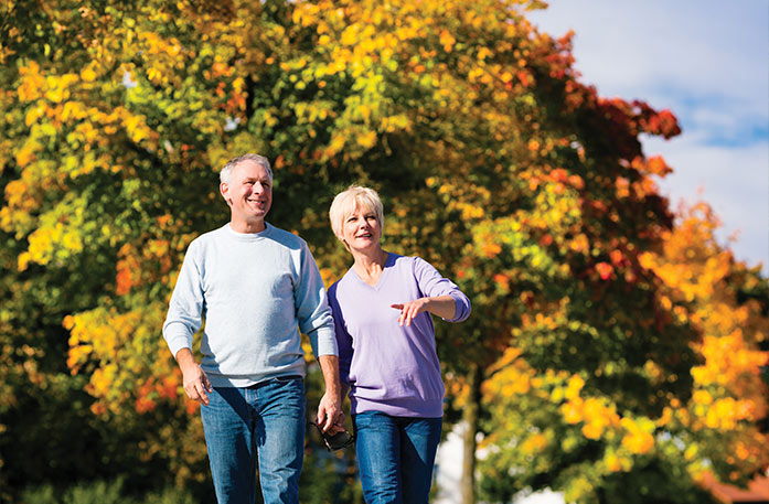 A couple walking and smiling in a park in Kingston during fall, with trees with orange, red, and green leaves in the background.