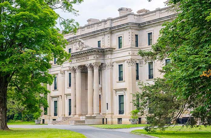 Vanderbilt Mansion National Historic Site in Hyde Park, a three-story, cream colored mansion with four tall pillars in front of the entryway stairs. There are two large trees outside of the mansion, and a perfectly manicured lawn.