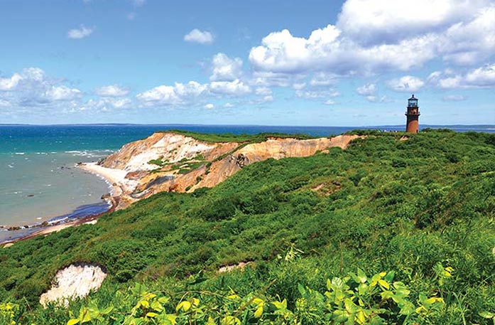 A large cliff beside the Massachusetts coastline in Martha's Vineyard. On top of the cliff among the greenery, there is a small, brown lighthouse, Gay Head Light.