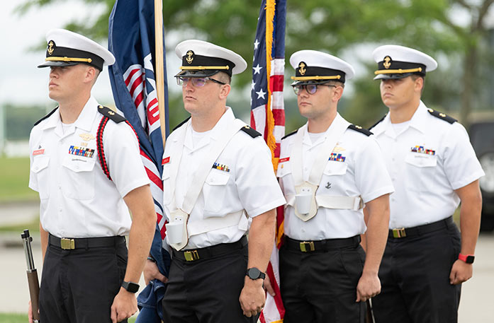 Four Massachusetts Maritime Academy cadets stand in a line wearing white uniforms with black pants and white and black sailors caps with a yellow anchor on the front. The two men in the middle are holding flags, one is an American Cruise Lines flag and the other is an American flag. 