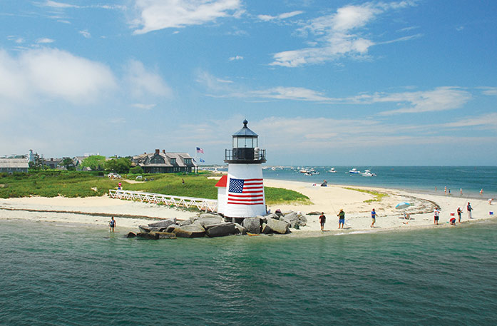 Brant Point Lighthouse, a small, white lighthouse on Nantucket Island with a large American flag on its side surrounded by rocks on the edge of the beach, along the water of Vineyard Sound. There is a ramp leading to the lighthouse from the grassy area behind the sand. People are fishing on the edge of the sand, and there are boats on the water and a large building in the middle of the island.