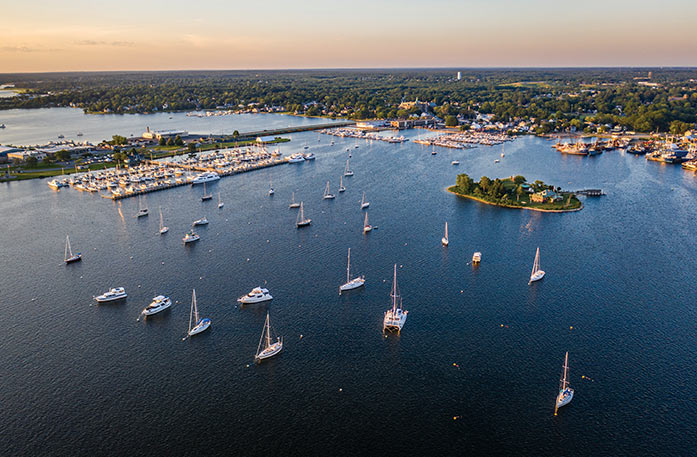 An aerial view of the bay with countless sailboats docked alongside an island and several sailing along the water with the city of New Bedford in the distance. 