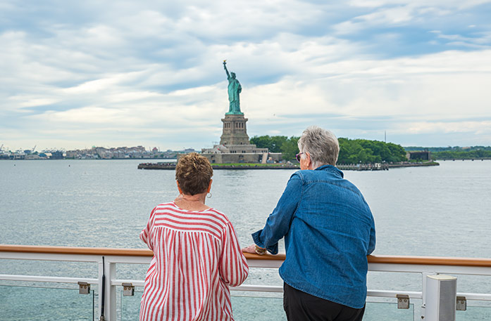 Two woman standing along the railing on the sun deck of an American Cruise Lines small cruise ship while looking out at the green Statue of Liberty in the near distance.