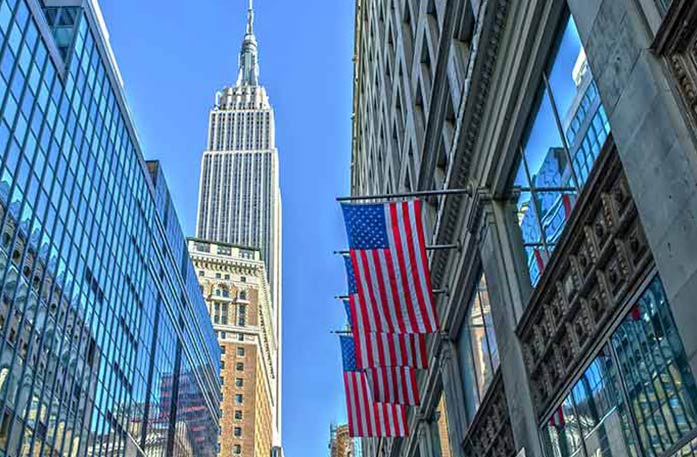 Four American flags hang from a building in New York, with the Empire State Building, a large skyscraper in the distance.