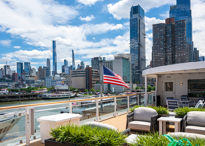 A view of the New York cityscape with countless skyscrapers and buildings from the sun deck of an American Cruise Lines small cruise ship. On the sun deck, there are lounge chairs and tables, as well as small planters. On the glass railing, there is an American flag.