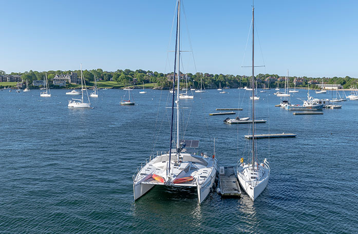 A large and a small sailboat are docked in the middle of Narragansett Bay in Newport, and several more are floating in the distance. On the shore, there are numerous trees, and a few large buildings.