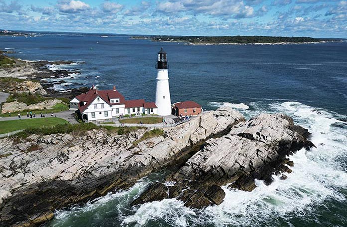 A birds-eye-view of Portland Head Light, a white lighthouse on top of a cliff alongside Casco Bay in Portland. There is a large white building with a dark red roof to the left of the lighthouse with several people walking nearby.