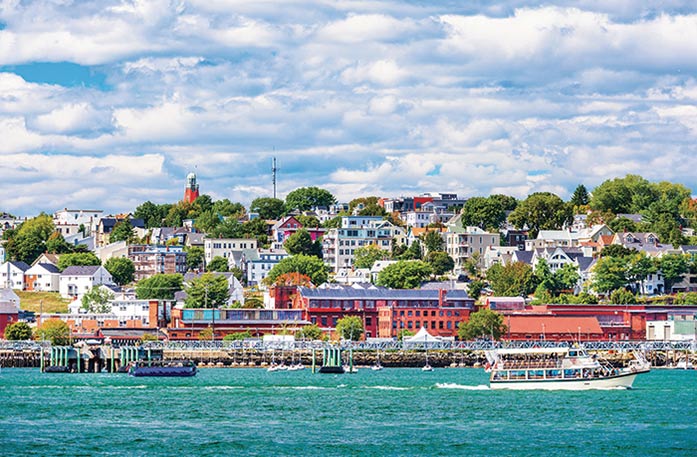 The Portland cityscape, with several colorful buildings on the shore with thick trees beside them, and boats sailing on Casco Bay in the foreground.