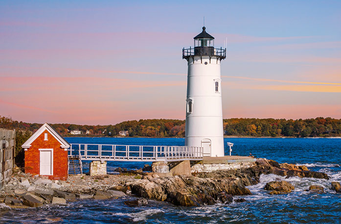 Portsmouth Harbor Lighthouse, a white lighthouse connected to a small red shed with a white door by a short bridge in Portsmouth. The lighthouse is on top of a large rock in the harbor, and the land on the other side of the water in the background is filled with fall foliage forest scenery under a pink sunset.