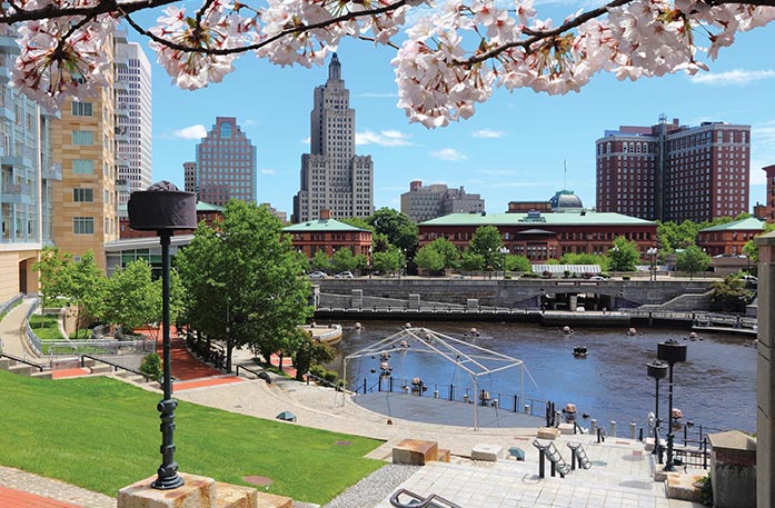 The heart of Downtown Providence, where tall buildings surround the Woonasquatucket River. Branches from a cherry blossom tree hang over the top of the foreground.