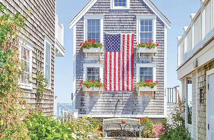 A tall and narrow grey house in Provincetown with four white windows, each with a flower box containing pink flowers, and a large American flag hanging on its side in the center. There is a house very close to each side, flowers and greenery alongside the houses, and a bench in front of the house.