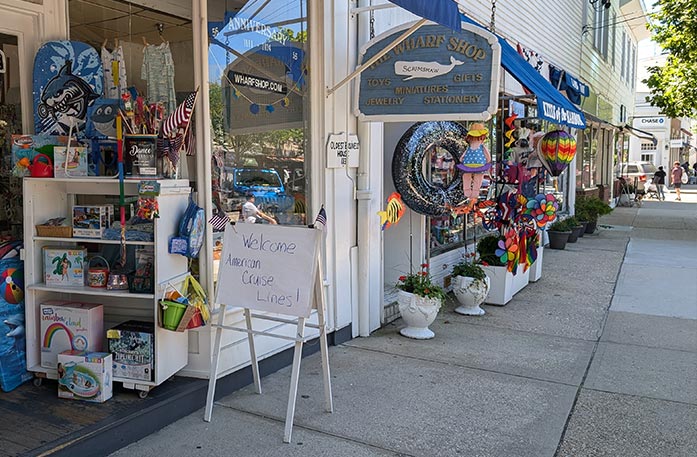 A line of shops along a sidewalk in Sag Harbor. The shop in focus is "The Wharf Shop", and has several items in shelves outside of the entryway, and a sign that says "Welcome American Cruise Lines!"